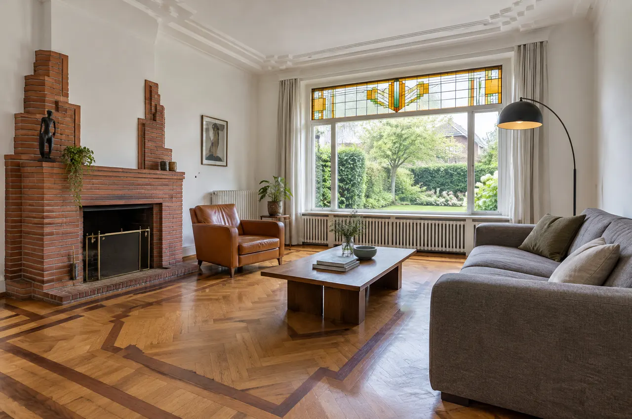 Salon de la villa : parquet géométrique d’origine, cheminée en brique, vitrail à motifs Art déco et fenêtre sur le jardin