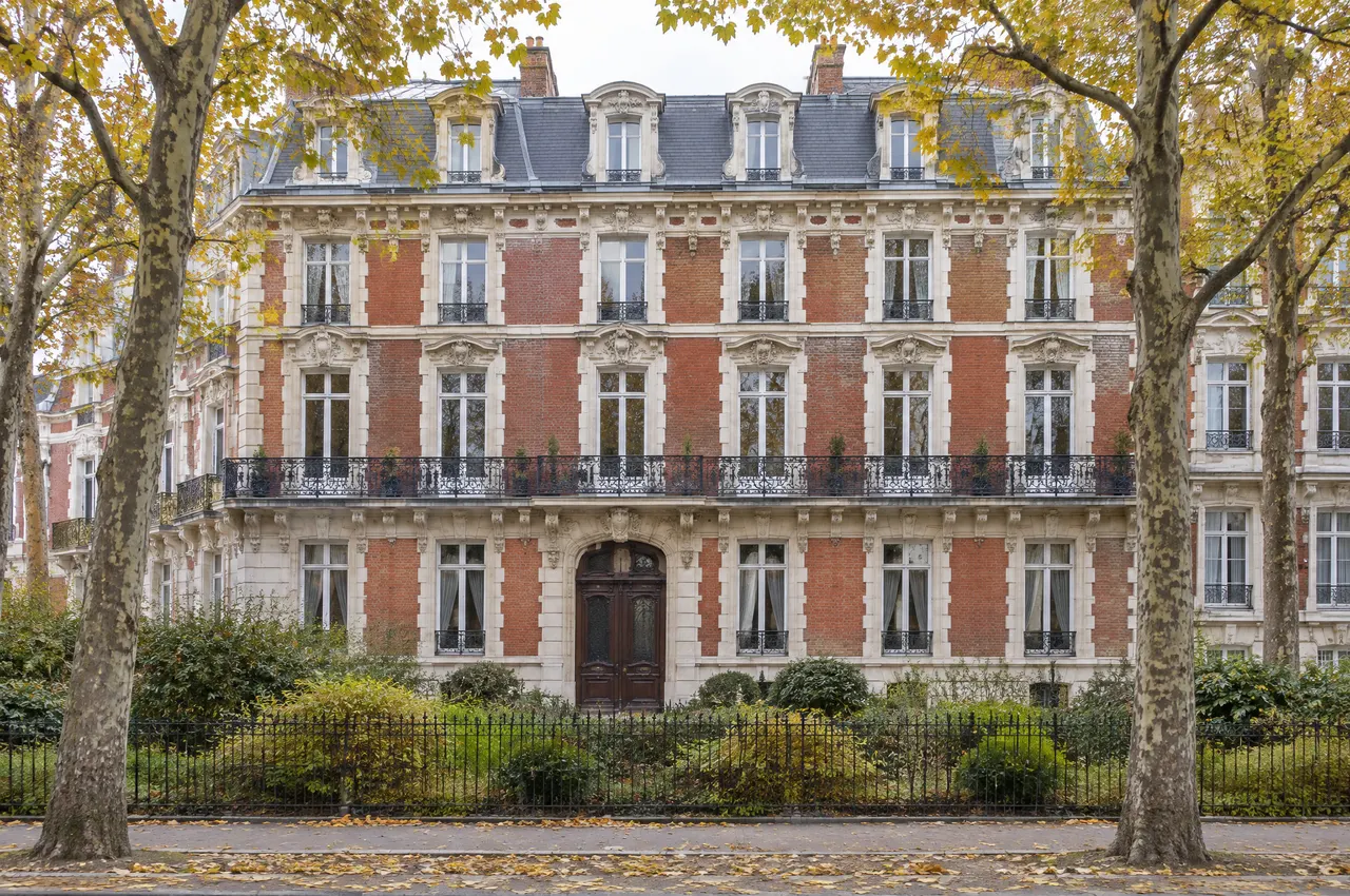 Immeuble 1900 en brique rouge et pierre blanche face au jardin, long balcon en fer forgé au deuxième étage, toit mansardé