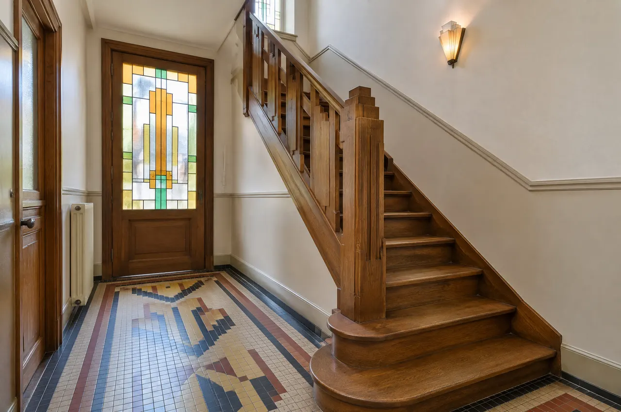 Entrée de la maison : carrelage d’origine à motifs géométriques, porte intérieure à vitraux et escalier en bois