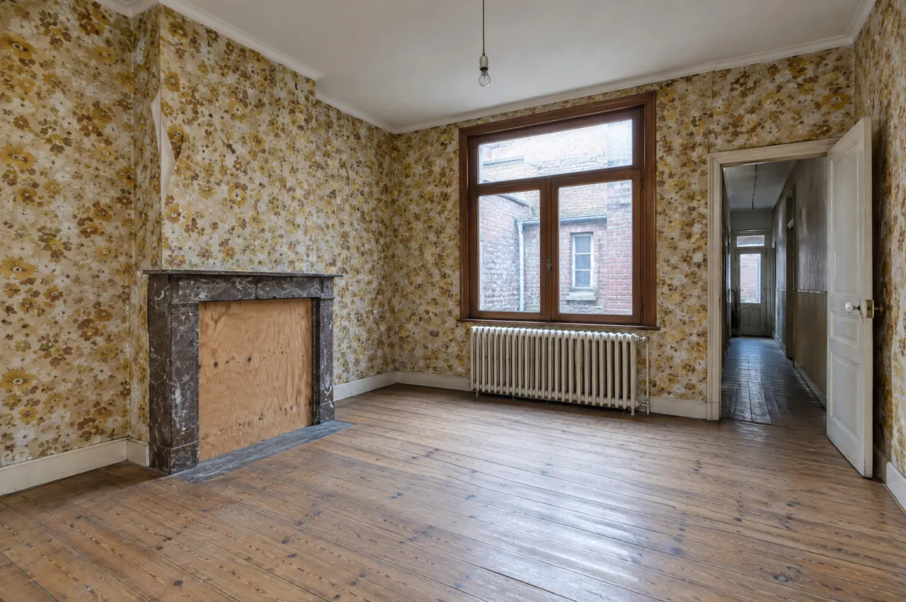 Pièce vide à rénover : papier peint défraîchi, cheminée condamnée, vieux parquet et fenêtre sur courette